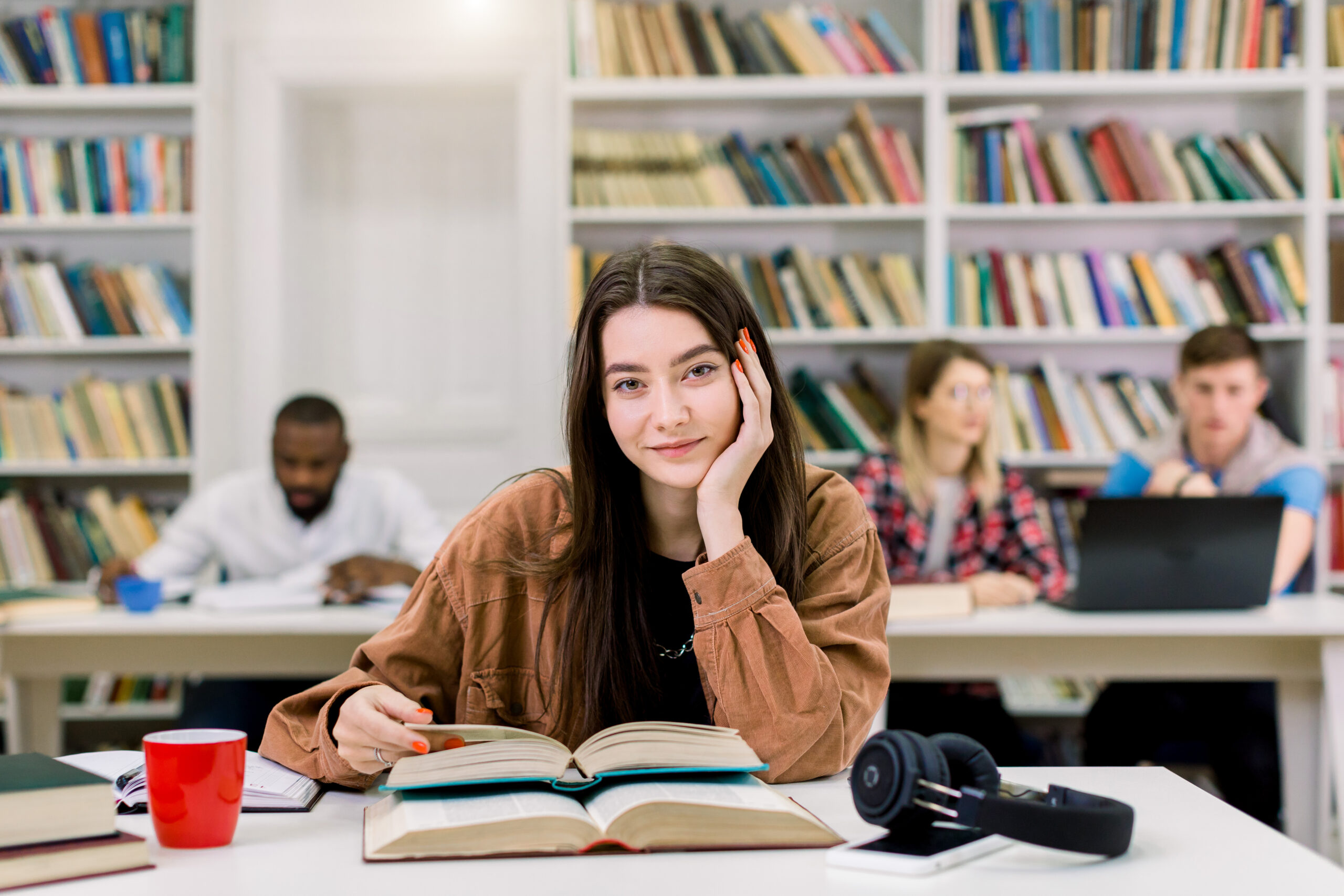 Two female student friends wearing protective face masks greeting each other bumping elbows while sitting at university library, healthcare concept, precautions, new normal
